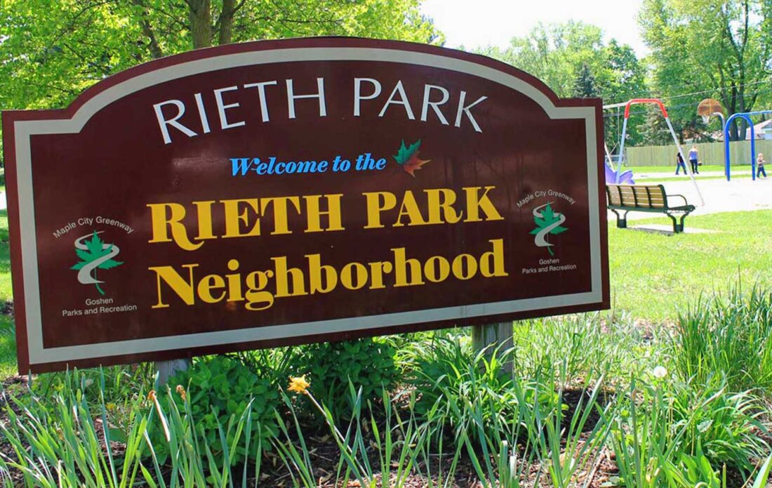 A photo of the Rieth Park welcome sign on a summer day with families enjoying the park's playground.